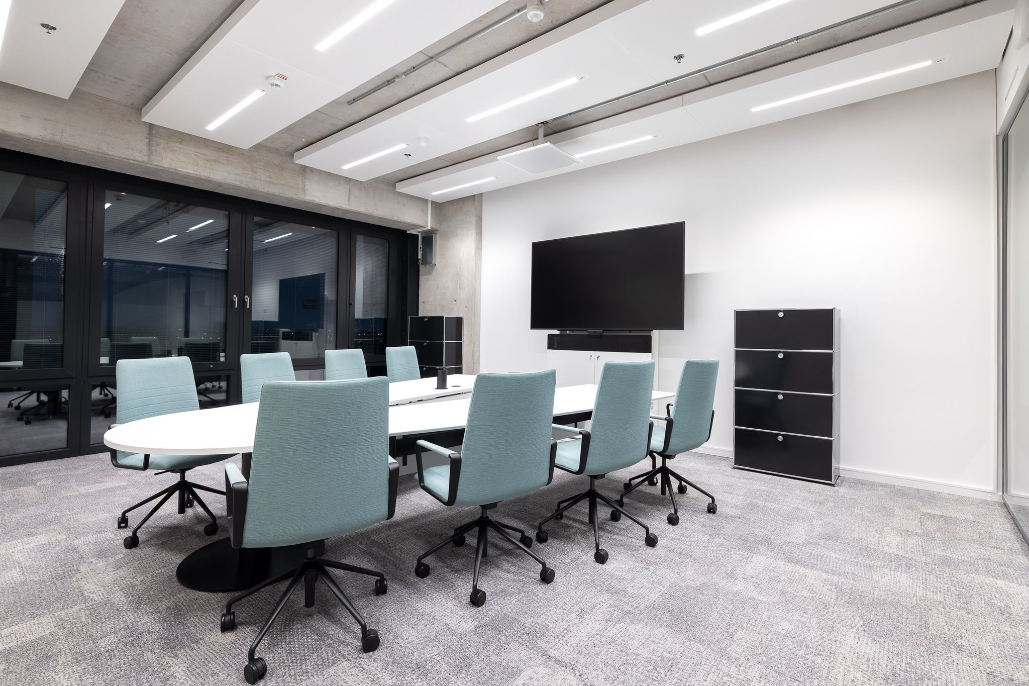 Kleines Besprechungszimmer mit Konferenztisch in Weiß mit 8 blauen Drehstühlen und Bildschirm an der Wand